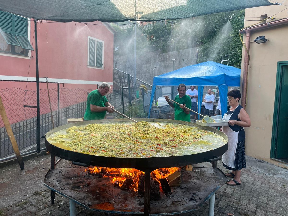 Volontari cucinano una grande paella in una padella enorme su fuoco a legna, nel cortile della sede durante una festa